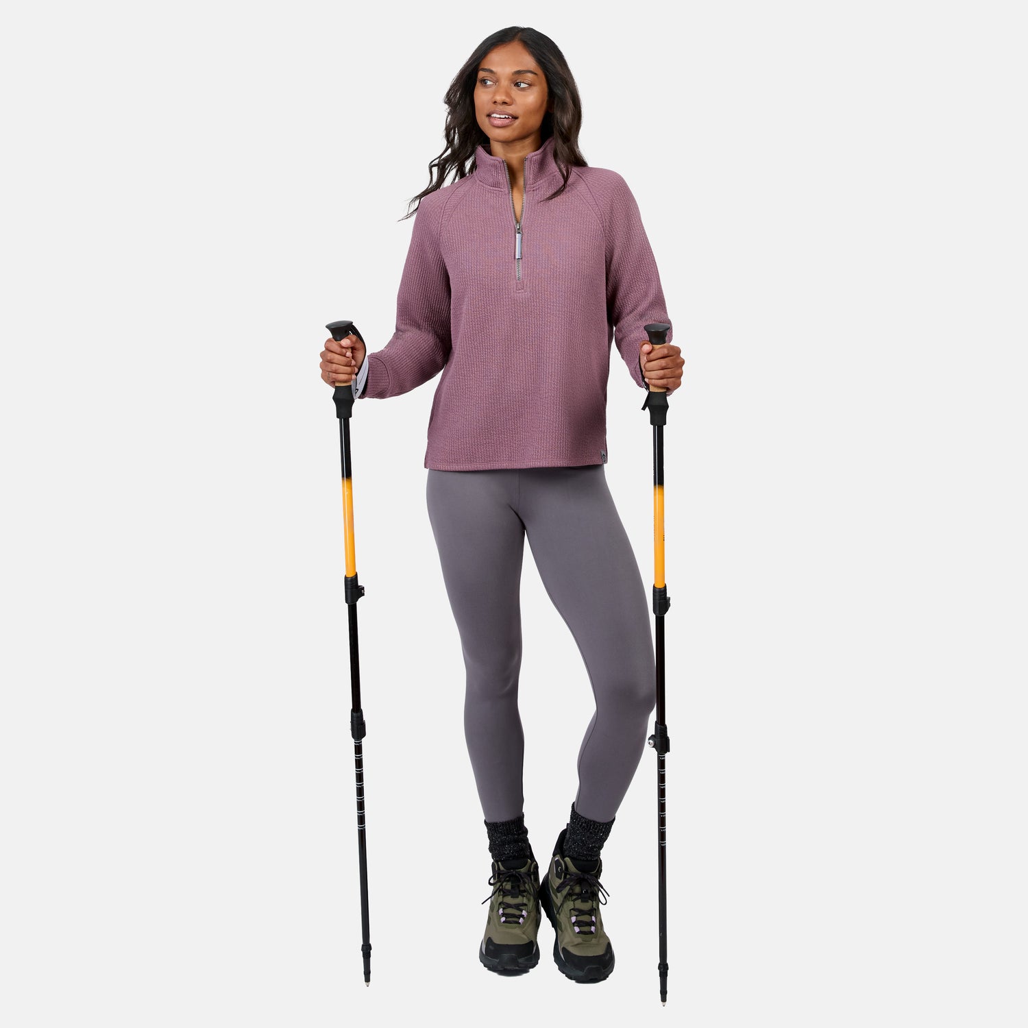 A woman stands with hiking poles, wearing the Free Country Women’s Ripple Knit Half Zip Top in mauve, gray leggings, black socks, and green hiking boots—breathable and perfect for casual layering on any outdoor adventure.