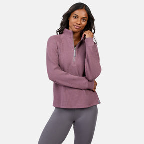 A woman wears the Free Country Women’s Ripple Knit Half Zip Top in mauve with gray leggings, standing against a white background, smiling with one hand in her hair—an ideal pick for casual layering.