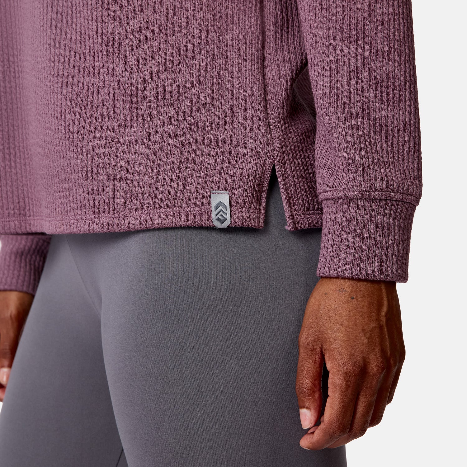 A woman wears the Free Country Women’s Ripple Knit Half Zip Top—a mauve, textured, breathable long-sleeve with a side tag—paired with fitted gray leggings, shown waist down with her right arm by her side.
