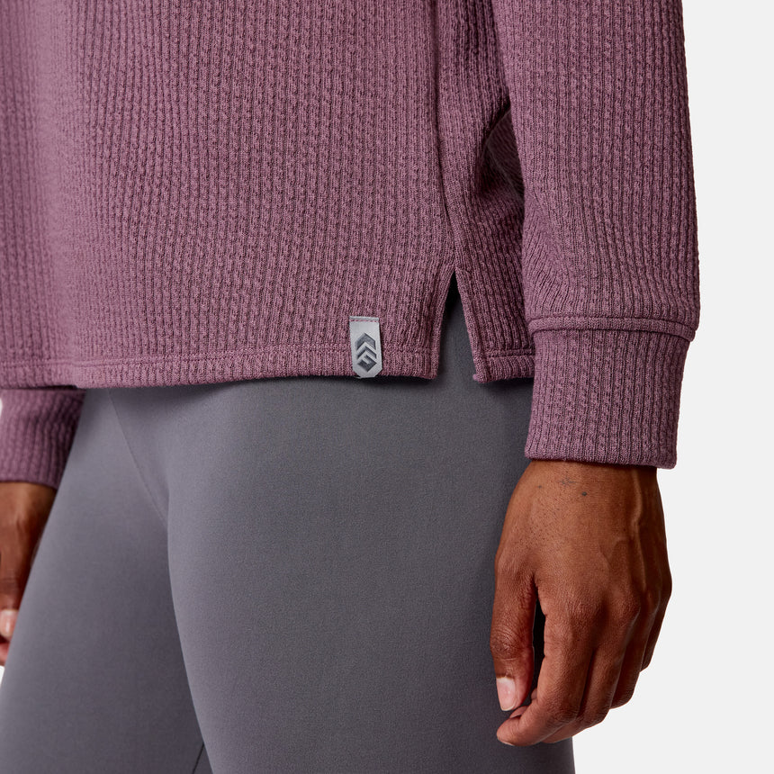 A woman wears the Free Country Women’s Ripple Knit Half Zip Top—a mauve, textured, breathable long-sleeve with a side tag—paired with fitted gray leggings, shown waist down with her right arm by her side.