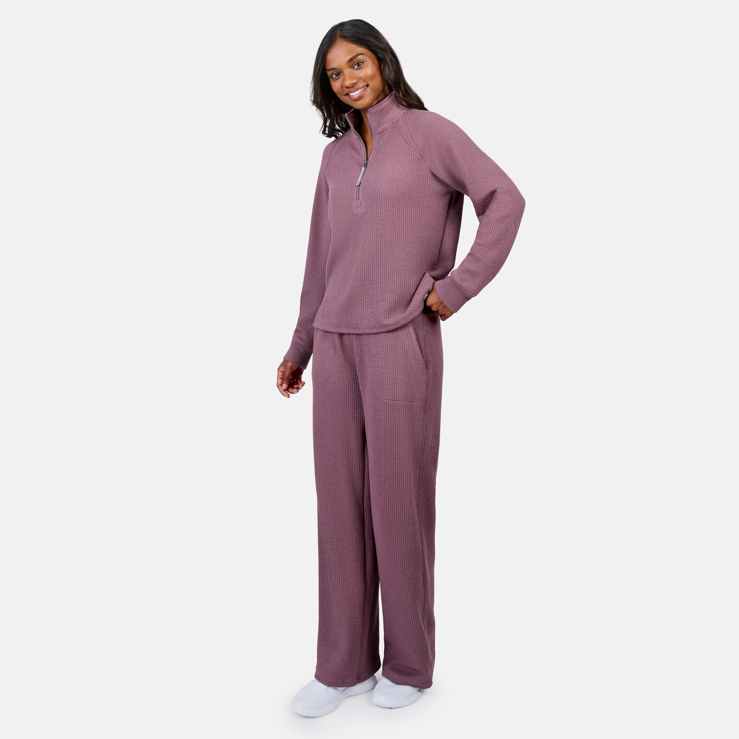 A woman smiles in Free Country’s Women’s Ripple Knit Half Zip Top and matching mauve pants, with her hand in her pocket. She pairs the breathable outfit with white slippers, perfect for layering against a plain white background.