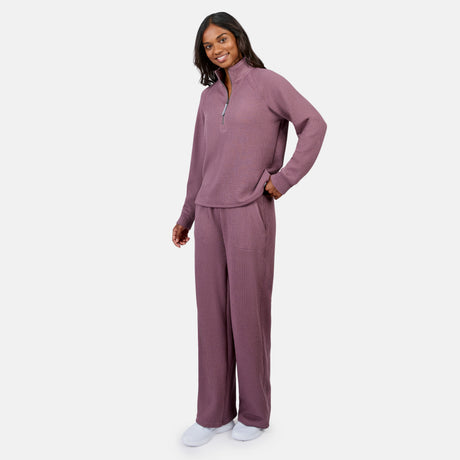 A woman smiles in Free Country’s Women’s Ripple Knit Half Zip Top and matching mauve pants, with her hand in her pocket. She pairs the breathable outfit with white slippers, perfect for layering against a plain white background.