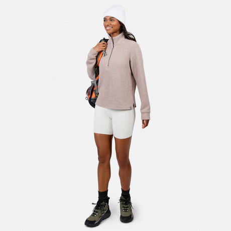 Woman in a Free Country Women’s Ripple Knit Half Zip Top, white bike shorts, beanie, and hiking boots smiles while holding a backpack—ideal for casual layering against a plain white background.