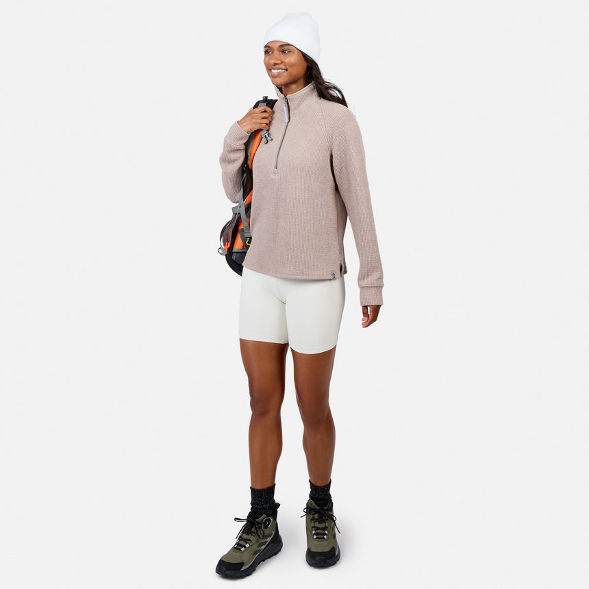 Woman in a Free Country Women’s Ripple Knit Half Zip Top, white bike shorts, beanie, and hiking boots smiles while holding a backpack—ideal for casual layering against a plain white background.