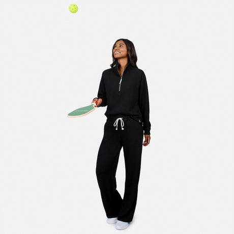 A woman in Free Country Women’s Ripple Knit Long Wide Leg Pant and white shoes smiles, holding a pickleball paddle and tossing a green pickleball, her pants providing comfort, all set against a plain white background.