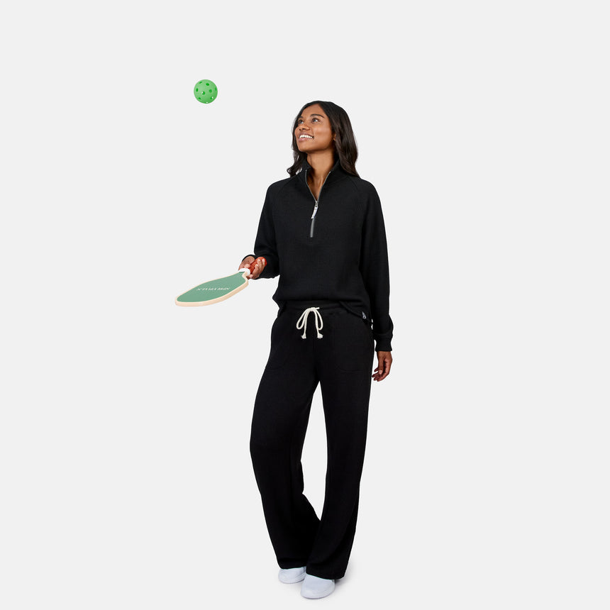 A woman in the Free Country Women’s Ripple Knit Half Zip Top and black pants smiles while balancing a green pickleball on a paddle, styled casually against a plain white background.