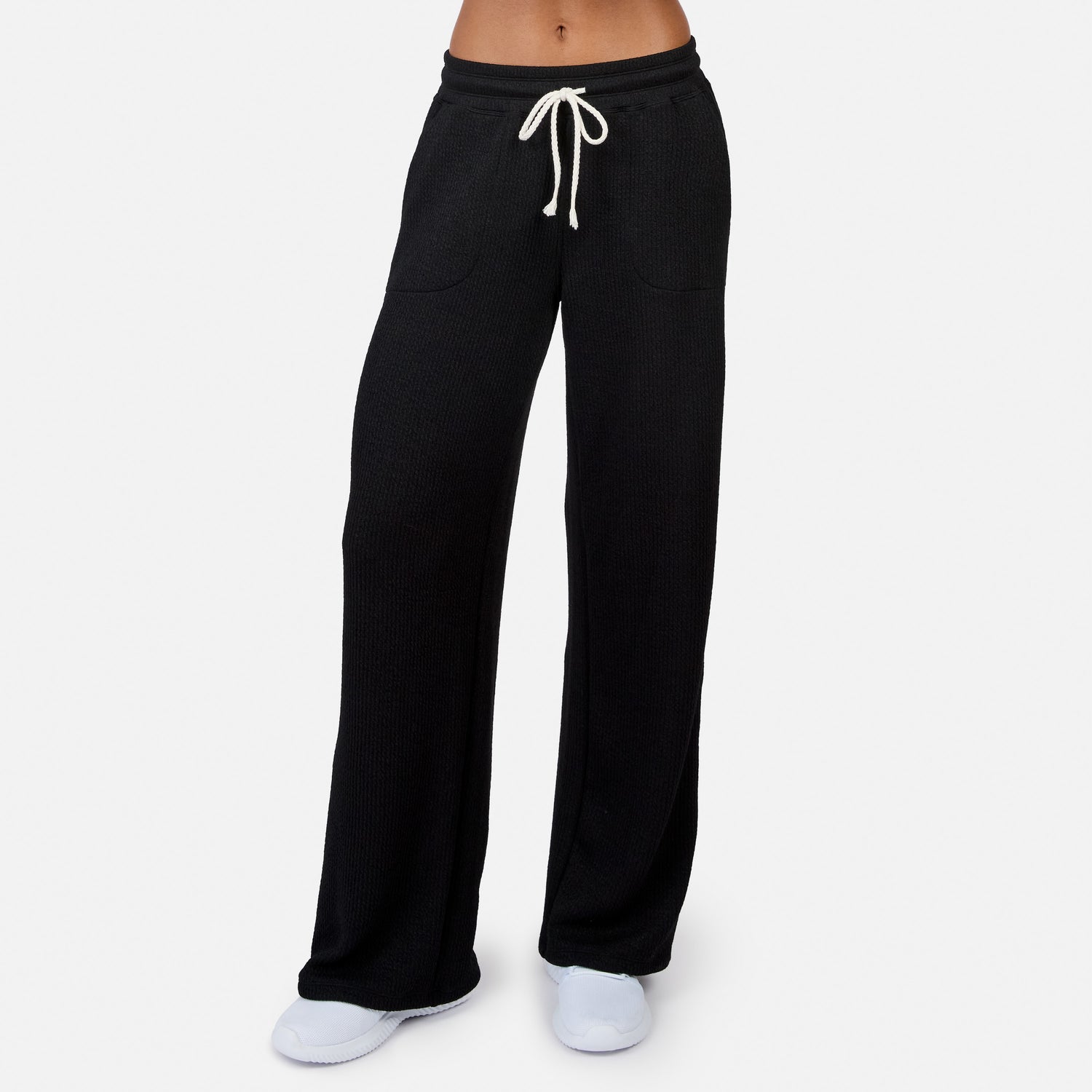 A person wears Free Country Women’s Ripple Knit Long Wide Leg Pant in black with a white drawstring, white shoes, and a bare midriff, standing against a plain white background.
