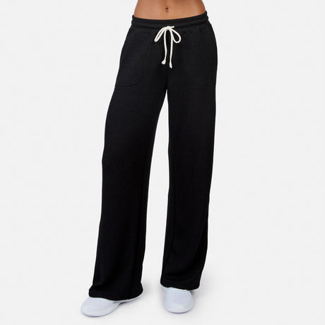 A person wears Free Country Women’s Ripple Knit Long Wide Leg Pant in black with a white drawstring, white shoes, and a bare midriff, standing against a plain white background.
