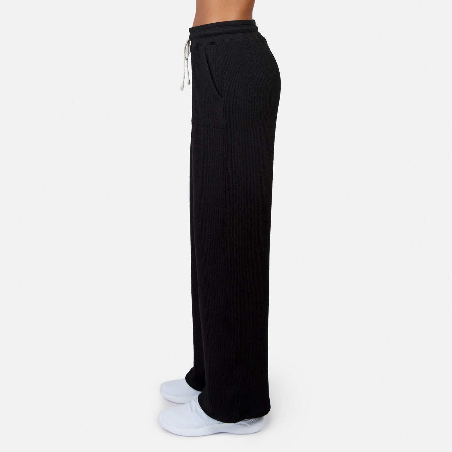 A woman wears Free Country Women’s Ripple Knit Long Wide Leg Pant in black with pockets and white sneakers, standing in profile from the chest down against a plain white background.