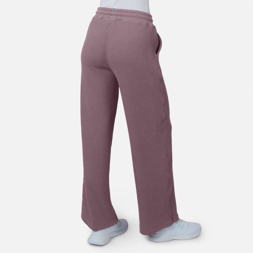 A person wears Free Country Women’s Ripple Knit Long Wide Leg Pants in maroon and white shoes, standing against a plain white background. The photo, taken from behind, showcases the pants’ relaxed fit and breathable ripple knit fabric.