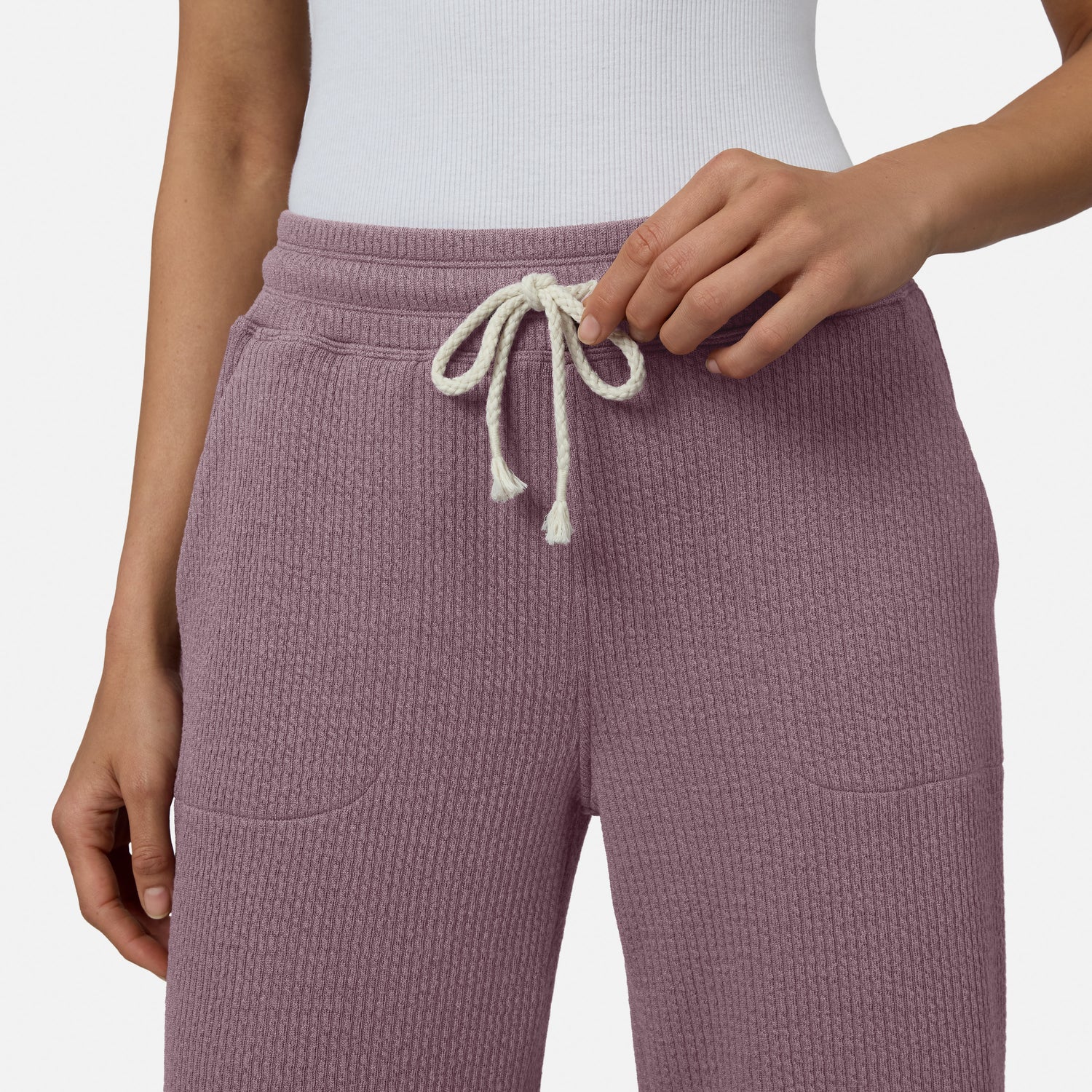 A person is shown from the waist down adjusting the drawstring of Free Country Women’s Ripple Knit Long Wide Leg Pants in purple, paired with a white ribbed top.