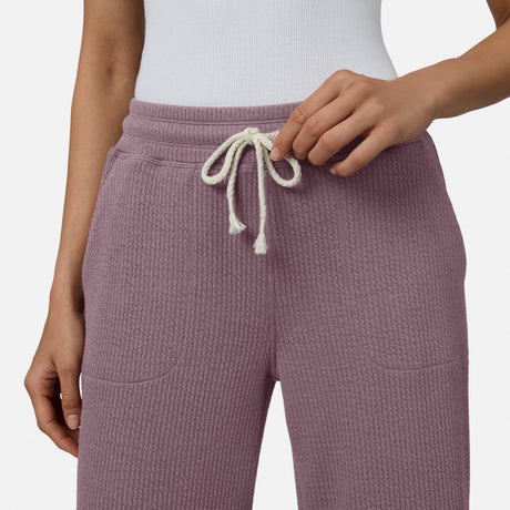 A person is shown from the waist down adjusting the drawstring of Free Country Women’s Ripple Knit Long Wide Leg Pants in purple, paired with a white ribbed top.