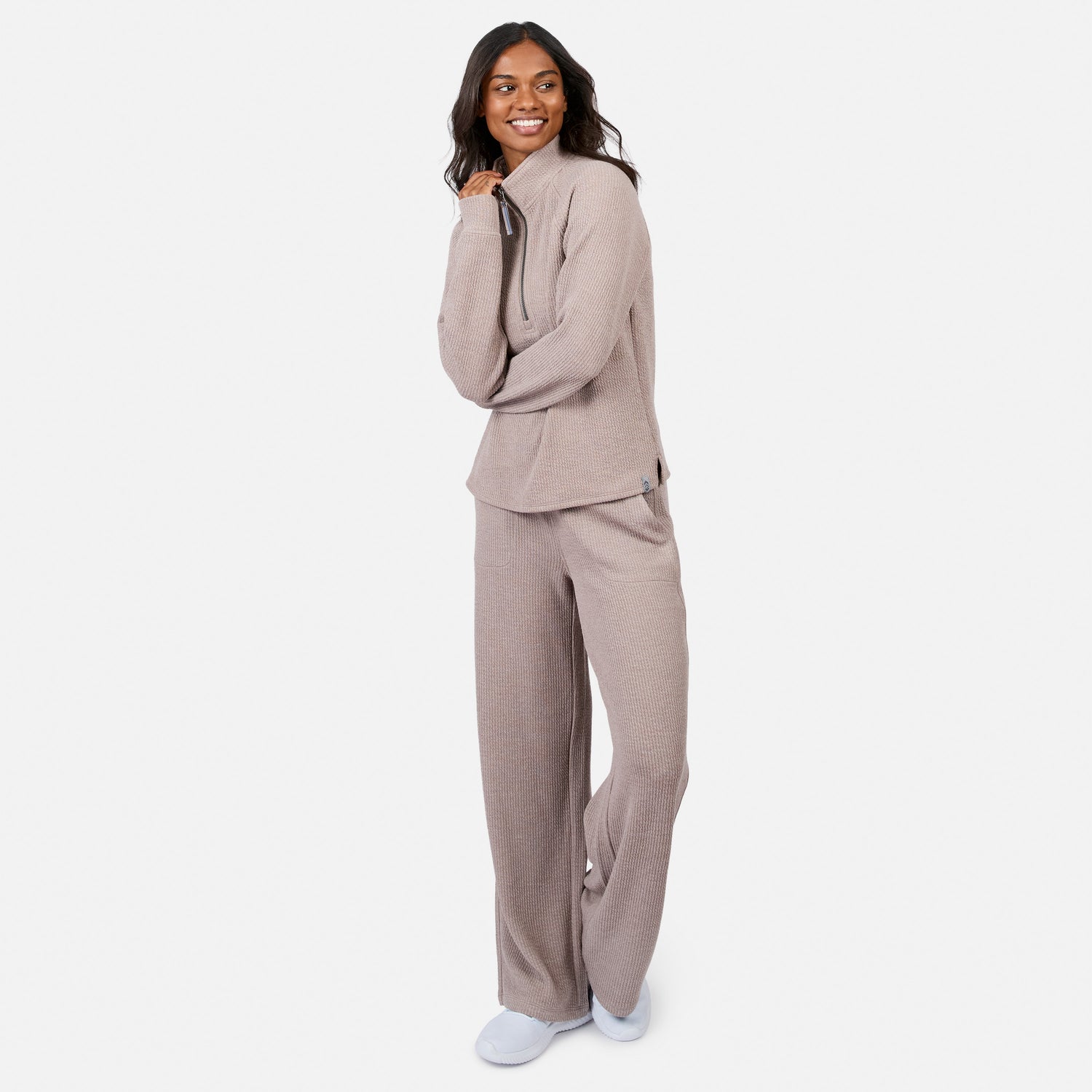 A woman smiles in the Free Country Women’s Ripple Knit Half Zip Top lounge set with wide-leg pants, both in light brown and breathable fabric, paired with white slip-on shoes, standing against a white background.