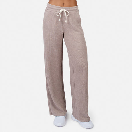 A person models the Free Country Women’s Ripple Knit Long Wide Leg Pant in taupe with a drawstring waist, paired with white socks and shoes, standing against a plain white background for a relaxed look.