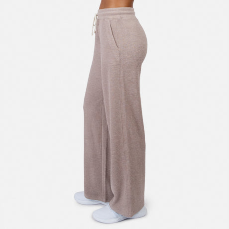 A woman stands in relaxed comfort from the side, wearing Free Country’s Women’s Ripple Knit Long Wide Leg Pant in beige with pockets and white slippers, against a plain white background.