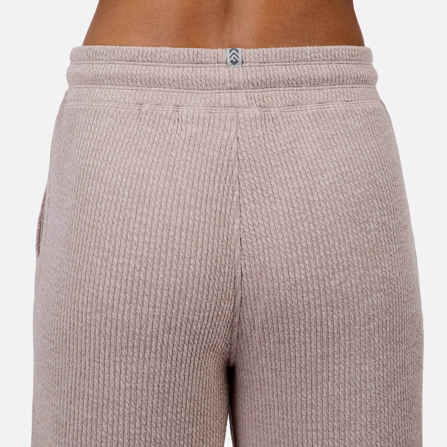 Close-up of the back of a person wearing Free Country Women’s Ripple Knit Long Wide Leg Pant in light brown, featuring ribbed fabric and a small gray and white logo centered at the waistband.