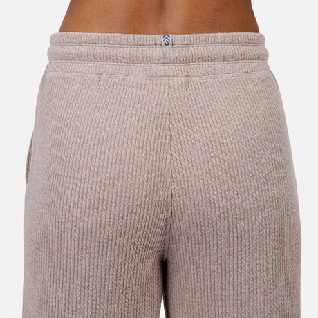 Close-up of the back of a person wearing Free Country Women’s Ripple Knit Long Wide Leg Pant in light brown, featuring ribbed fabric and a small gray and white logo centered at the waistband.