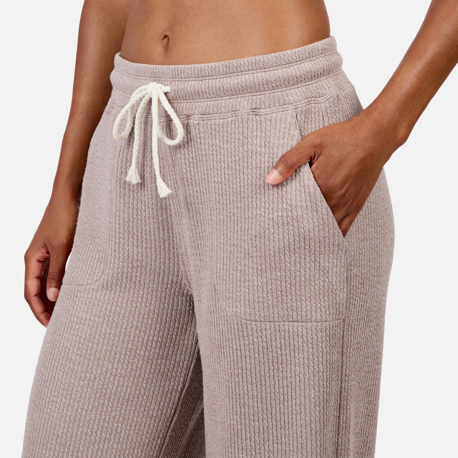 A model shown from the waist down wears Free Country Women’s Ripple Knit Long Wide Leg Pant in light brown, featuring a breathable textured fabric and drawstring waist, with their right hand in the pocket against a plain white background.