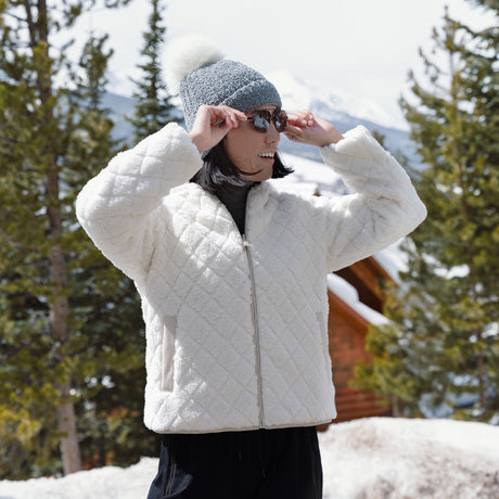 Person wearing a white quilted jacket and gray beanie in a snowy landscape with trees and a cabin.