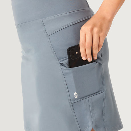 A person places a black smartphone into the side pocket of a light blue Free Country Women's Free 2 Explore Hybrid Skort, showcasing its four-way stretch and comfort; only the hand, lower torso, and skort are visible against a plain background.