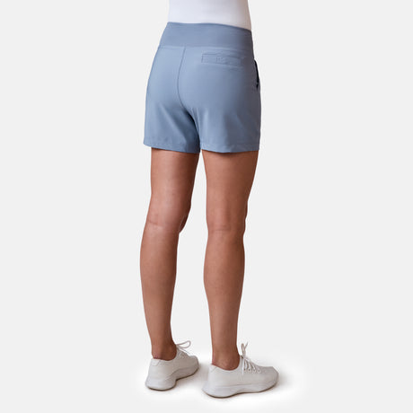 Facing away, a person wears Free Country's Women's Free 2 Explore Hybrid Short in light blue with four-way stretch, a fitted waistband, and back pocket, paired with a tucked-in white shirt and sneakers against a plain light backdrop.