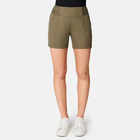 Against a plain white background, only the lower half of a person is visible. They're wearing Free Country's Women's Free 2 Explore Hybrid Short in olive green, paired with a black top and white sneakers.
