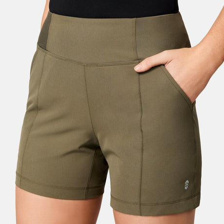 A person wears Free Country Women's Free 2 Explore Hybrid Shorts in olive green, with one hand in the pocket. Visible stitching, moisture-wicking fabric, and a small logo on the left leg are shown from waist to mid-thigh.