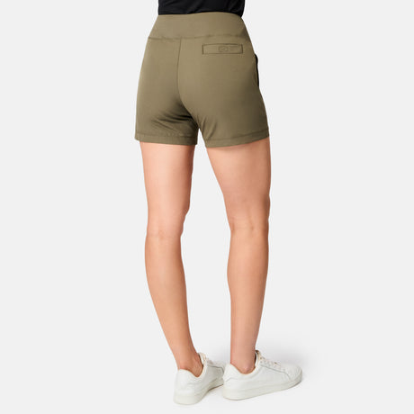 A person is standing with their back to the camera against a white background, wearing Free Country Women's Free 2 Explore Hybrid Shorts in olive green, paired with a black top and white sneakers.