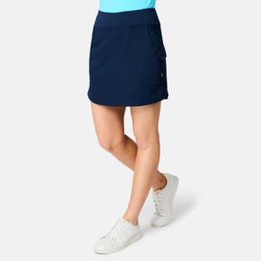 A woman wearing the Free Country Women's Free 2 Explore Hybrid Skort in navy blue with side pockets, a light blue top, and white sneakers stands against a plain white background.