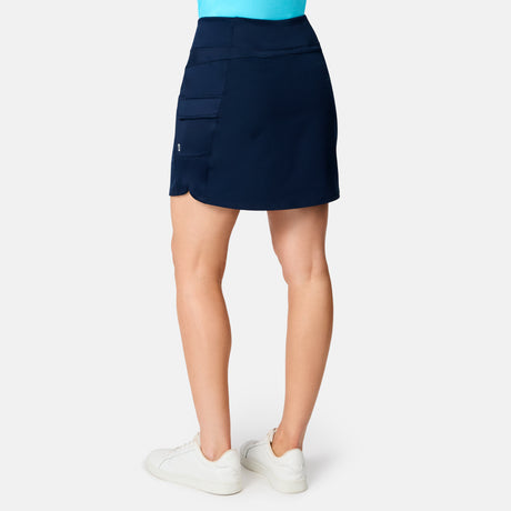 A woman is shown from the back wearing the Free Country Women's Free 2 Explore Hybrid Skort in navy blue, featuring side pockets, a slightly curved hem, and four-way stretch fabric. She pairs it with white sneakers against a plain white background.