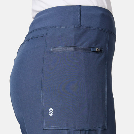 Close-up of a person wearing Free Country Women's Trail to Town Short in blue stretch knit, featuring a zippered back pocket and small white geometric logo on the lower part. The background is plain and light-colored.