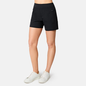 A woman wears Free Country's Women's Trail to Town Short in black stretch knit with side pockets and a small left-side logo, paired with white sneakers, standing against a plain white background from waist to feet.