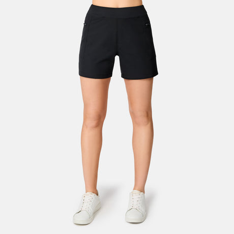 A person models the Free Country Women's Trail to Town Short in black with white sneakers, standing against a plain white background. The cropped photo focuses on the legs and feet, highlighting the shorts' stretch knit design.