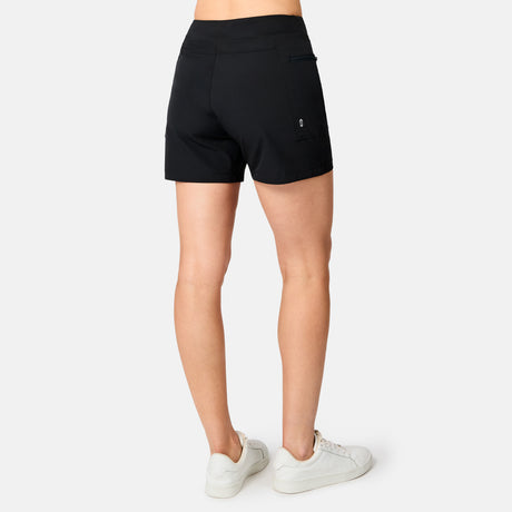 A woman stands facing away, wearing Free Country Women's Trail to Town Shorts in black with a back cargo zipper pocket and white sneakers, on a plain white background.