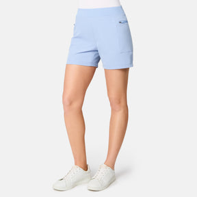 A person wears the Free Country Women's Trail to Town Short in light blue, featuring moisture-wicking fabric and a zippered pocket, paired with white sneakers against a plain white background. Only the lower half of the body is visible.