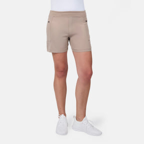 A person models the Free Country Women's Trail to Town Short in beige, featuring stretch knit fabric and cargo zipper pockets, paired with white sneakers, standing against a plain white background with their upper body out of frame.