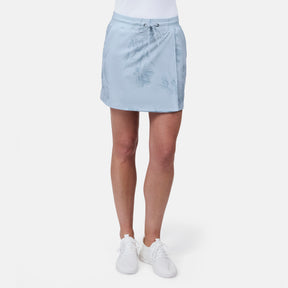 A person is wearing a light blue Free Country Women's Trail to Town Skort with subtle floral patterns, paired with a white top and white sneakers, standing against a plain white background.