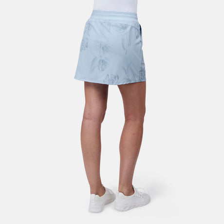 A person stands against a plain white background wearing the Free Country Women's Trail to Town Skort in light blue with a subtle leaf pattern, paired with white sneakers and a white top.