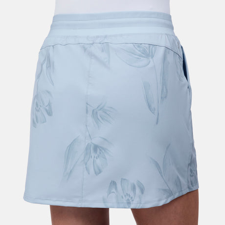 A woman models the Free Country Women's Trail to Town Skort in light blue with subtle floral patterns, featuring four-way stretch fabric, side pockets, and a smooth waistband. The background is plain white.