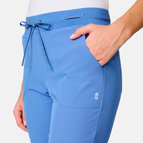 A person wears the Free Country Women's Get Out There Ruch Up Pant in light blue, featuring quick-dry fabric, elastic waistband with drawstring, front pockets (one hand tucked in), and a small logo on the thigh.