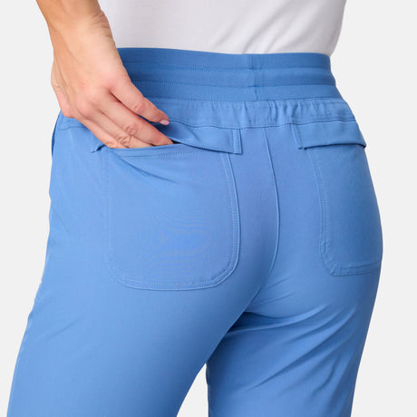 A person in light blue Women's Get Out There Ruch Up Pants by Free Country with a white shirt places their left hand into the back pocket, drawing attention to the waistband and pocket details.