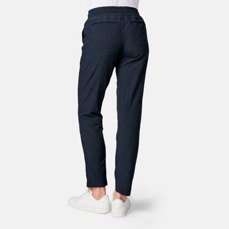 Model wears the Women's Get Out There Ruch Up Pant by Free Country in navy blue and white sneakers, seen from the back against a light gray background. These tapered, quick-dry pants offer moisture-wicking fabric and practical back pockets.