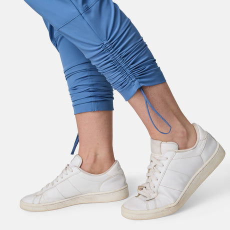 A close-up of a person wearing Free Country Women's Get Out There Ruch Up Pant in blue with white sneakers, standing on tiptoe against a plain white background.