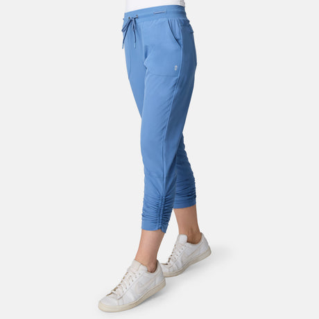 A woman wears the Free Country Women's Get Out There Ruch Up Pant in light blue with ruched cuffs and white sneakers, standing against a plain white background.