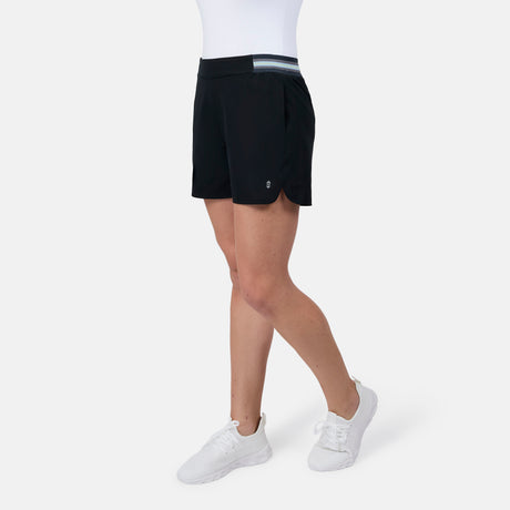 A person wearing Free Country Women's Explorer Shorts in black with a striped waistband, paired with a white top and white sneakers, stands against a plain white background. Only the lower body is visible.