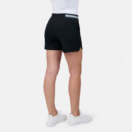 A woman in a white top, Free Country Women's Explorer Short with a striped waistband, and white sneakers stands facing away against a plain white background.