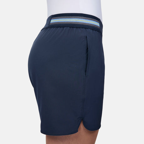 A woman wears the Free Country Women's Explorer Short in navy blue, featuring a blue and white striped elastic waistband, visible pocket, and curved hem. These moisture-wicking shorts provide comfort for any adventure.
