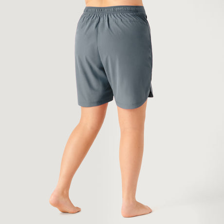 A woman stands barefoot on a light background, seen from the back, wearing Free Country's Women's Plus Size Bermuda Board Short II—loose, gray knee-length shorts with an elastic waistband and quick-drying fabric.