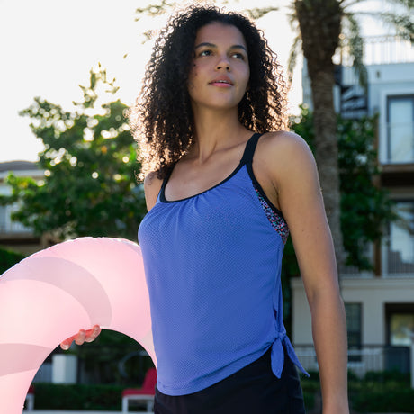 A woman with curly hair wears the Free Country Women's Textured Mesh Side Tie Blouson Tankini Top in blue while holding a pink and white inflatable pool float outdoors, with palm trees and a building in the background.