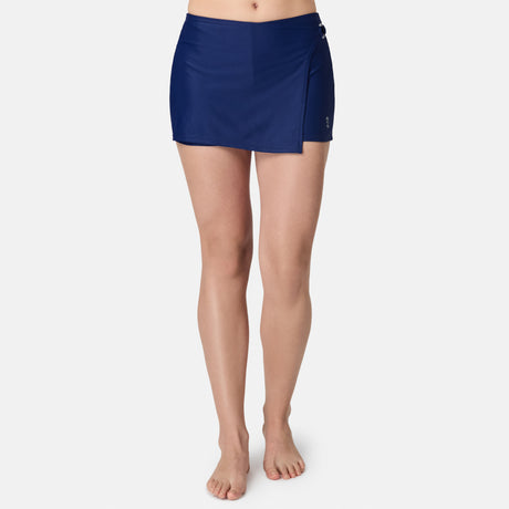 A barefoot woman wears the Free Country Women's Fully Lined Swim Skort, featuring a navy blue wrap design, against a plain white background.